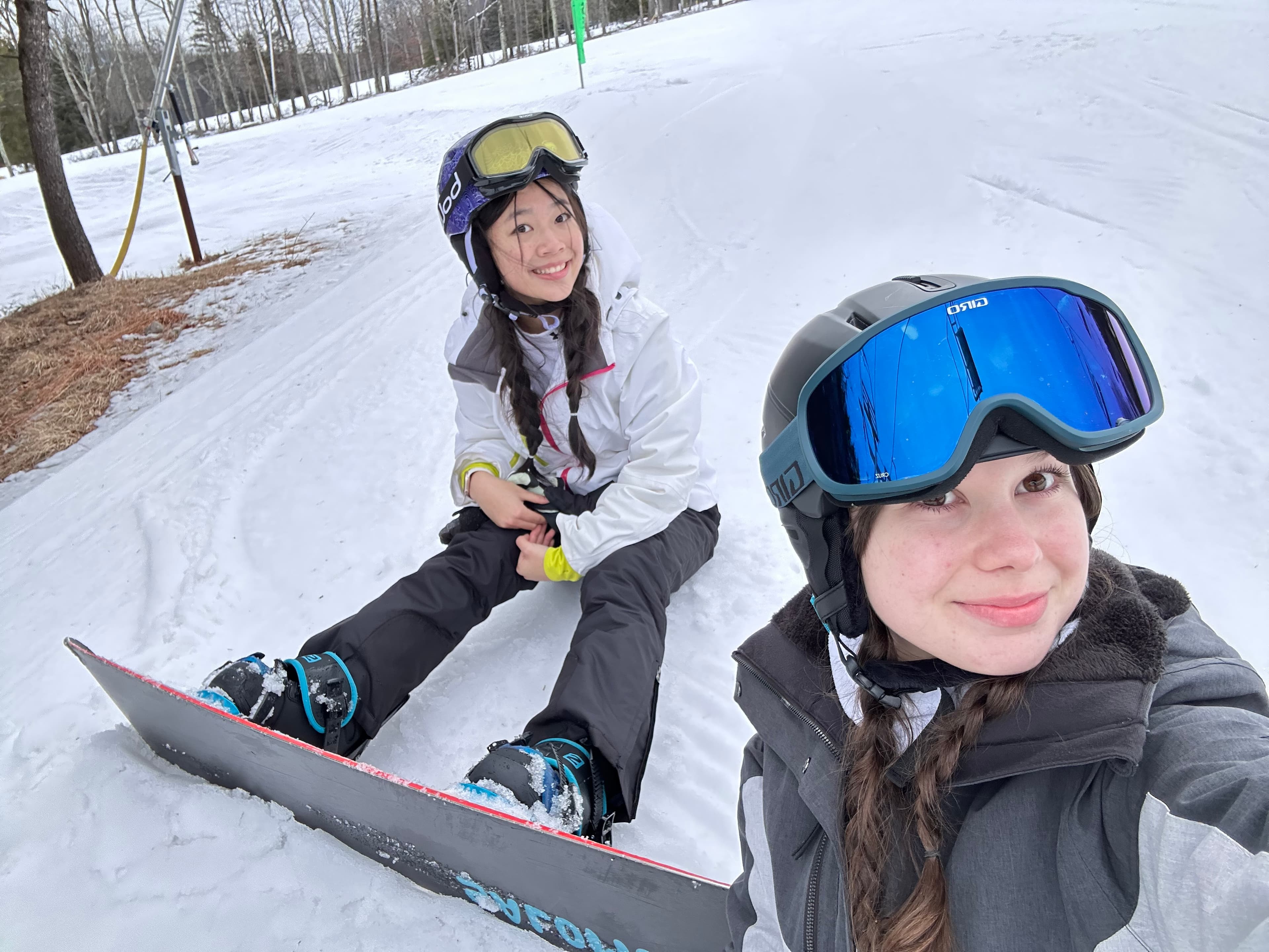 Doris sitting on a snowy slope in a helmet and goggles, next to a friend